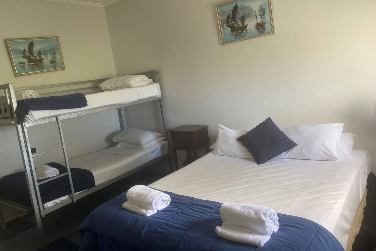 A hotel room with a bunk bed, a single bed, and a small table. There are two paintings on the walls.