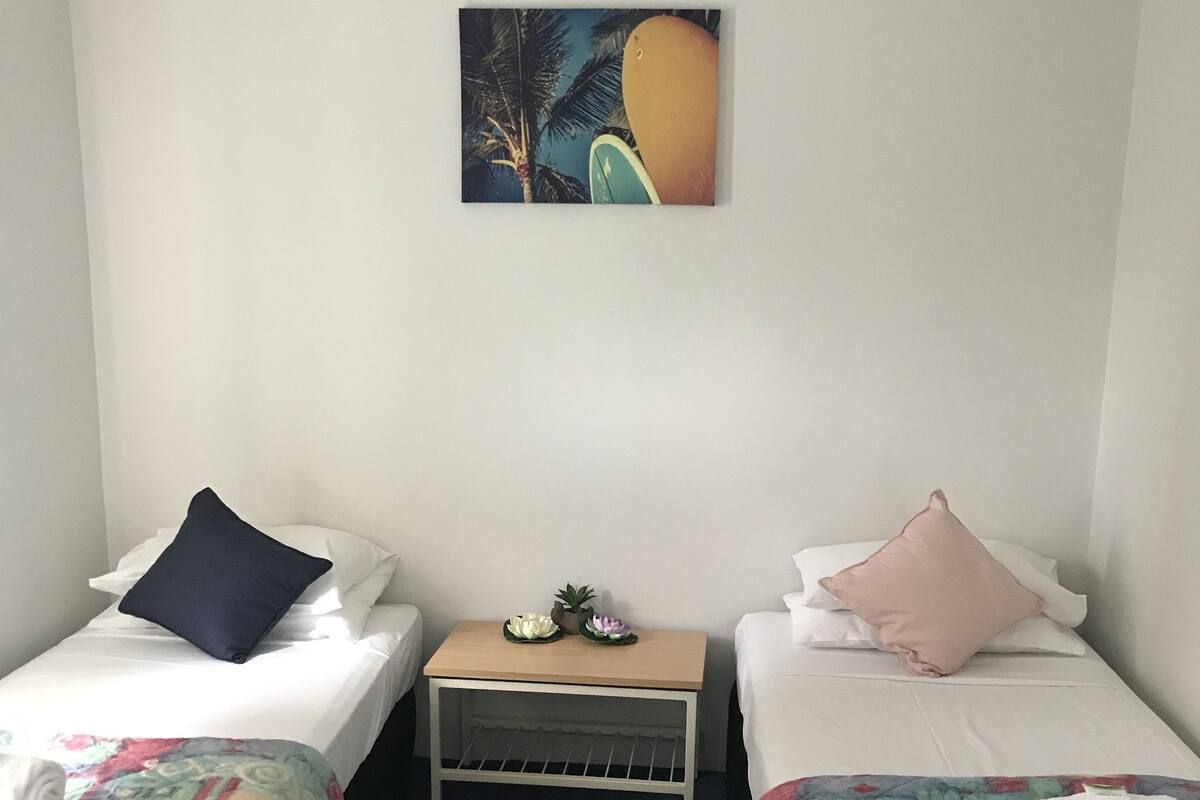 A room with two single beds, each with a colorful quilt and a pillow. There is a small table with a plant and a decorative item between the beds. A painting is hung on the wall above the table.
