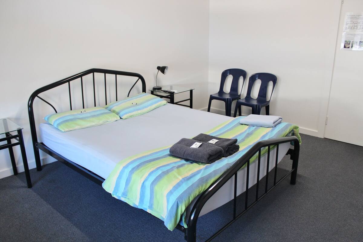A single bed with a striped bedspread, a pair of chairs, a small table with a lamp, and a wall-mounted bulletin board.