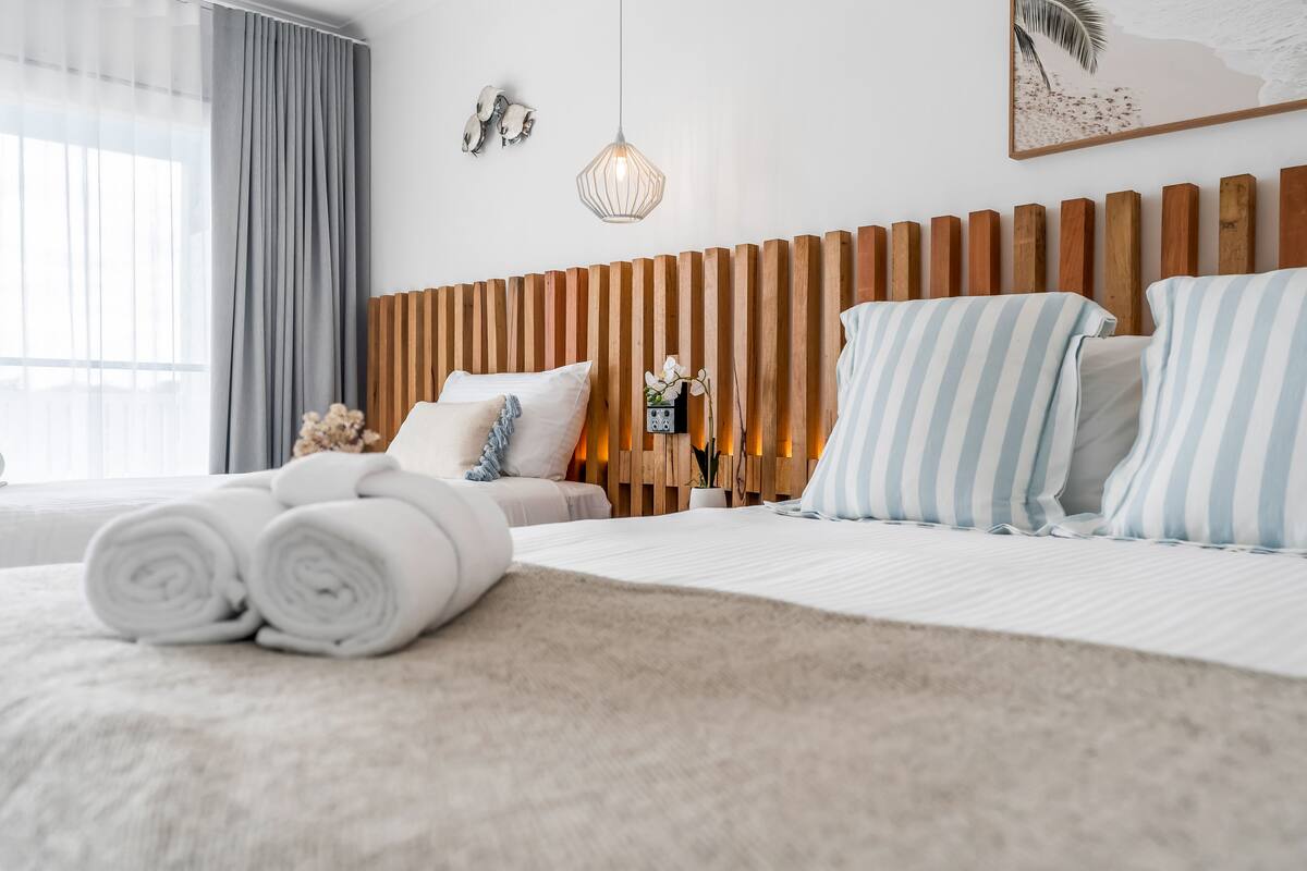 A modern hotel room with a wooden headboard, a large bed with striped pillows, a view of the city through sheer curtains, and a decorative wall piece.
