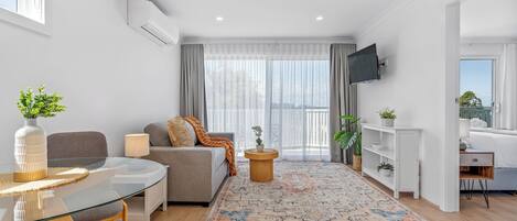 A modern living room with a glass dining table, grey sofa, and a large window with sheer curtains.