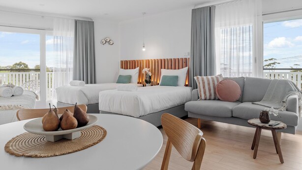 A modern hotel room with a large bed, a sofa, a dining table with a fruit bowl, and a balcony with a view.
