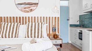 A bedroom with a wooden headboard, a bedside table with a lamp, and a view of a kitchen with a refrigerator and cabinets.