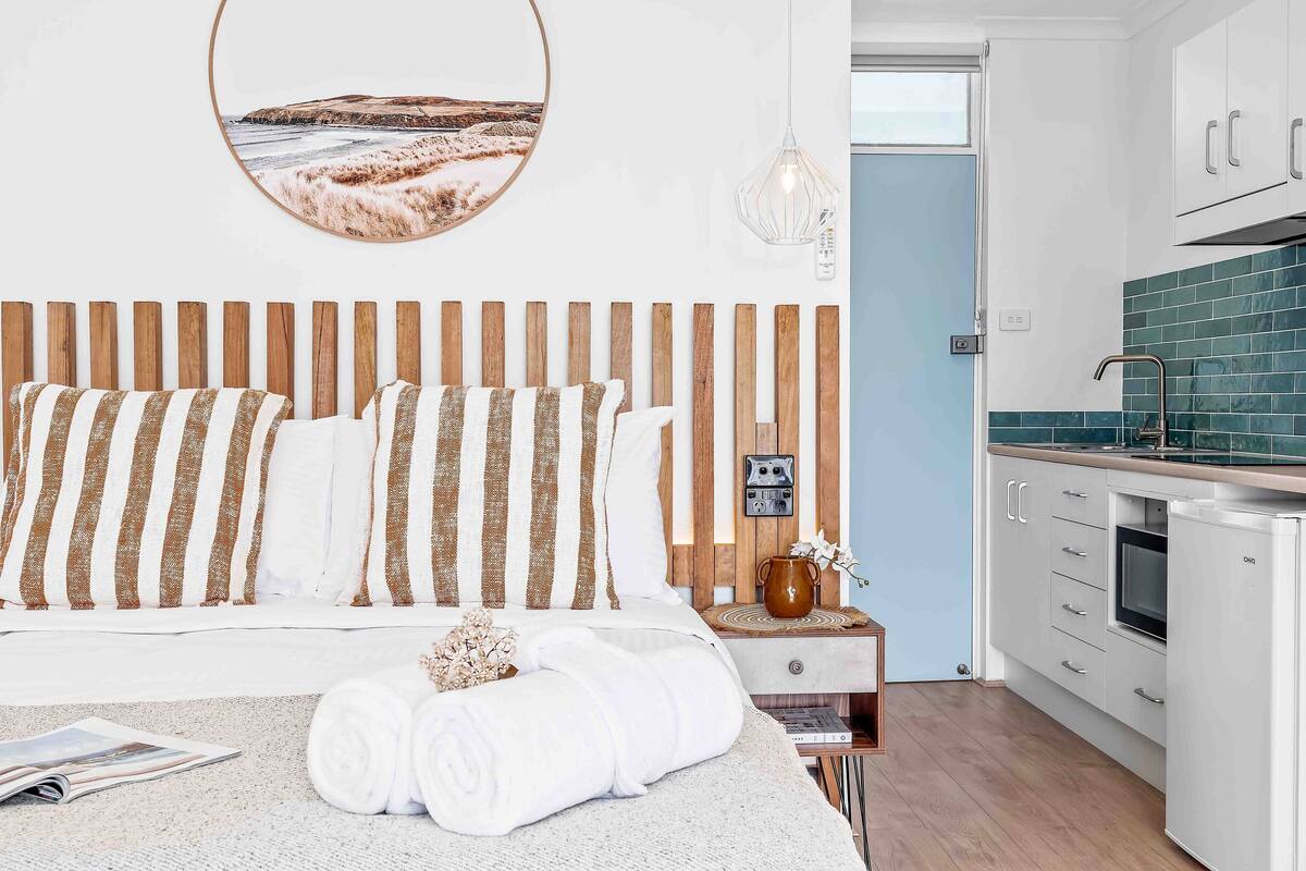 A bedroom with a wooden headboard, a bedside table with a lamp, and a view of a kitchen with a refrigerator and cabinets.