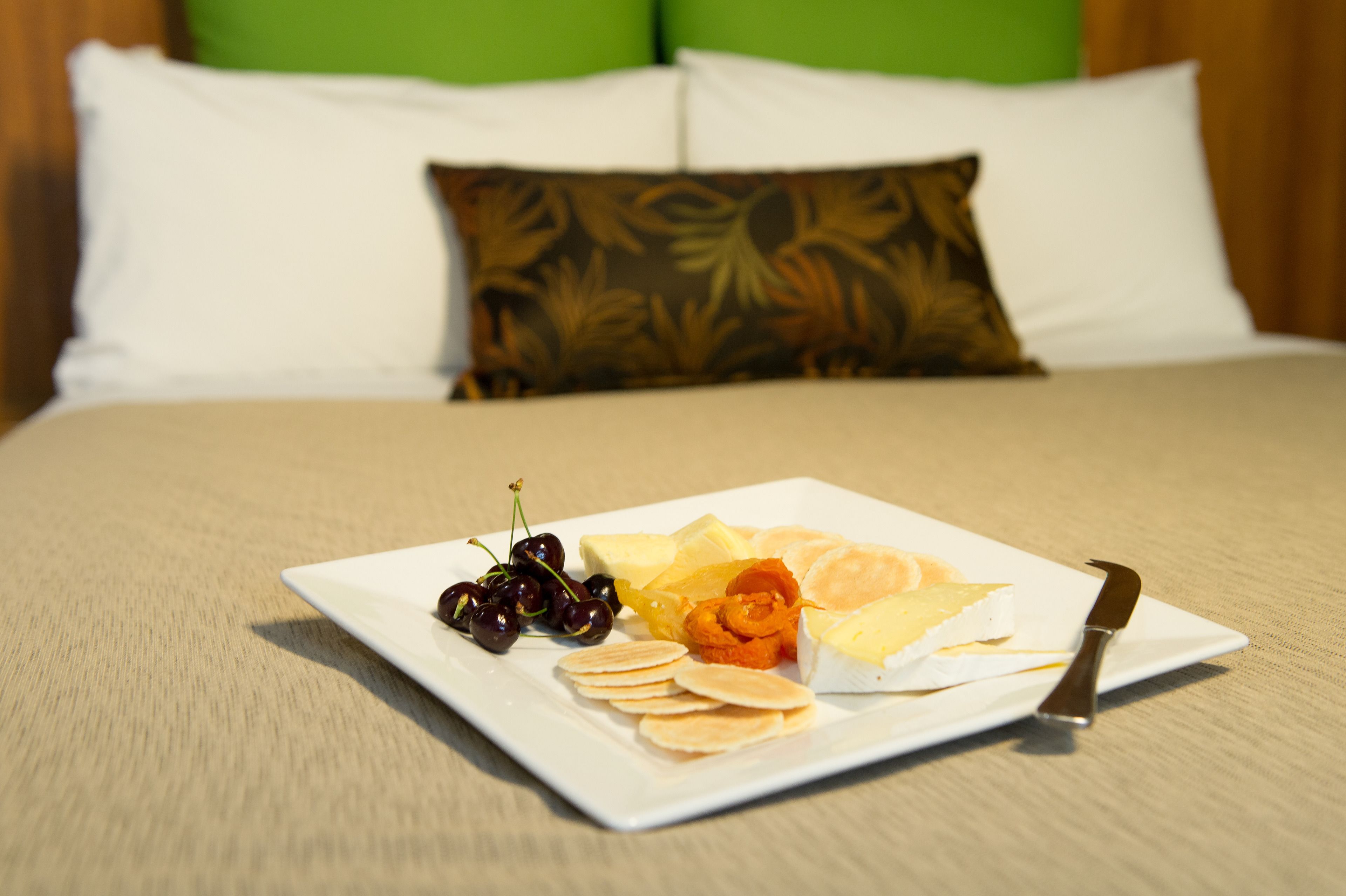 A neatly arranged breakfast plate with various fruits and pastries on a white plate, accompanied by a knife, set on a bed with a beige bedspread and green pillows in the background.