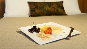 A neatly arranged breakfast plate with various fruits and pastries on a white plate, accompanied by a knife, set on a bed with a beige bedspread and green pillows in the background.