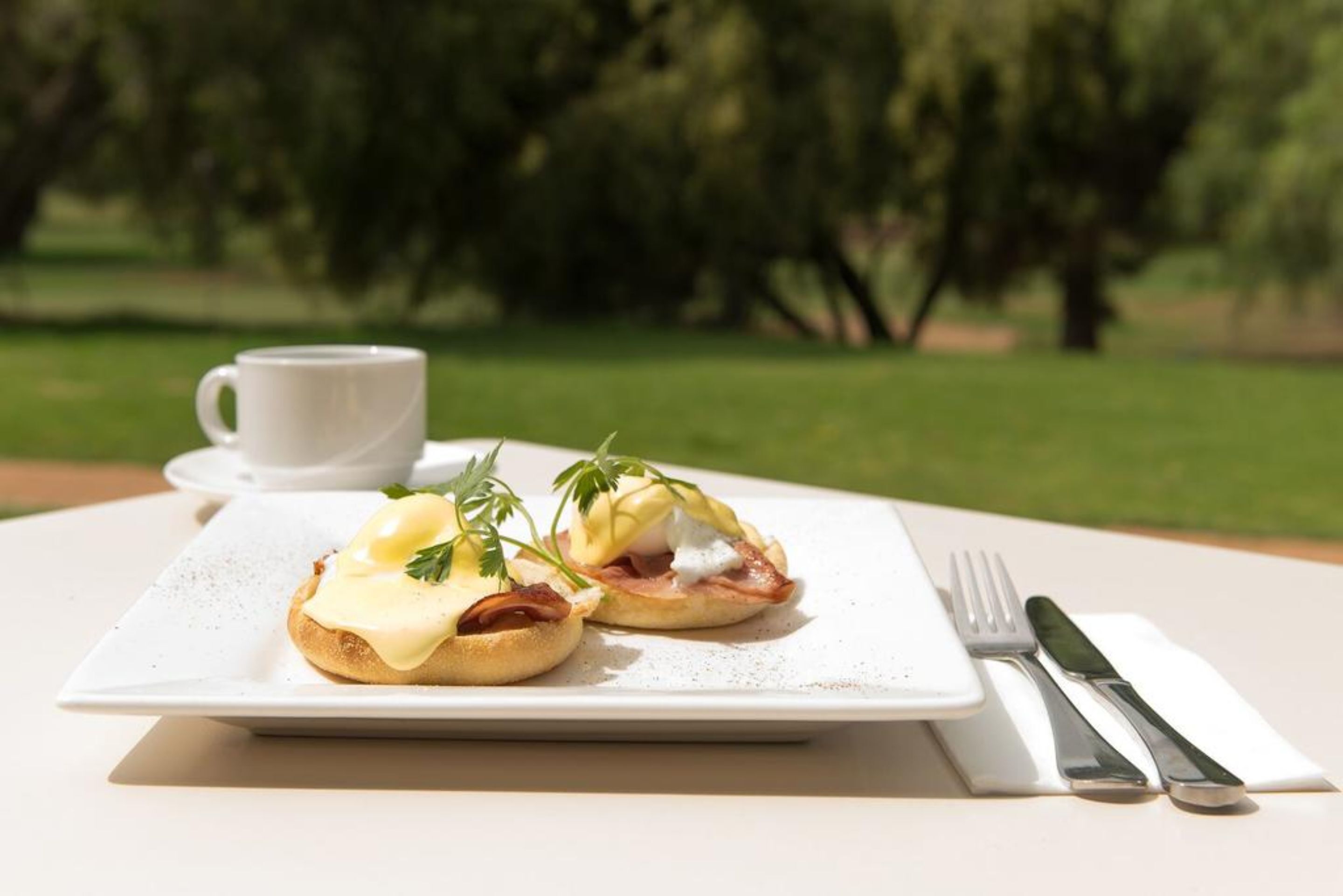 Une table dressée pour le petit-déjeuner, avec deux œufs Bénédicte sur une assiette blanche, une tasse blanche et des couverts sur une table blanche.