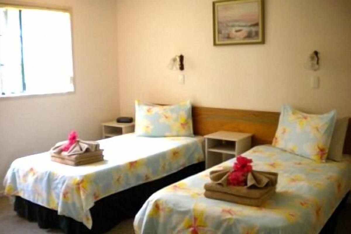 Habitación de hotel con dos camas individuales, cada una con ropa de cama floral y una mesita de noche pequeña. Hay una ventana a la izquierda y un cuadro en la pared sobre las camas.