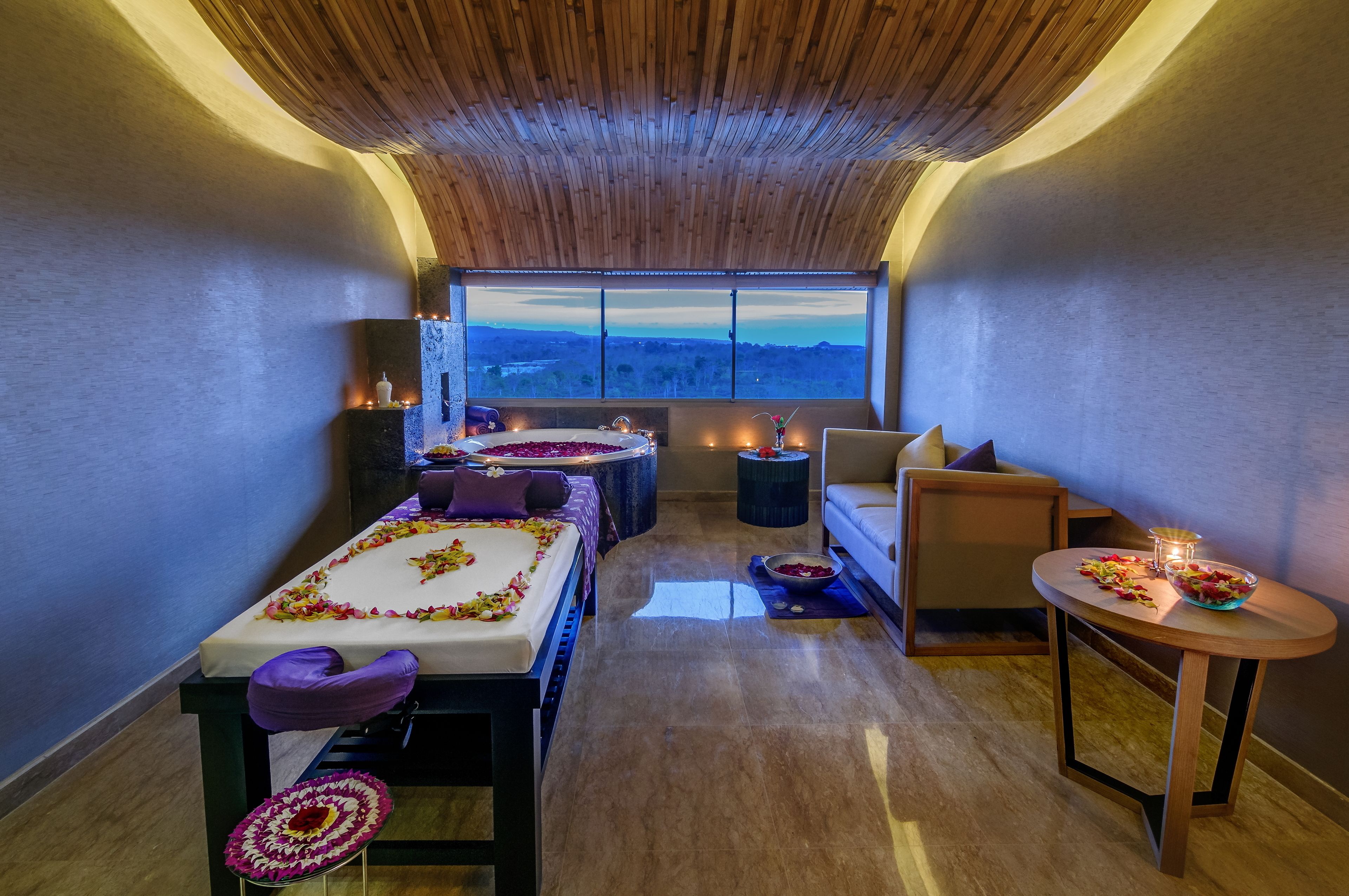 Une salle de spa équipée d’une table de massage, d’un petit canapé, d’une table ronde avec des bougies et offrant une vue sur le paysage grâce à une grande fenêtre.