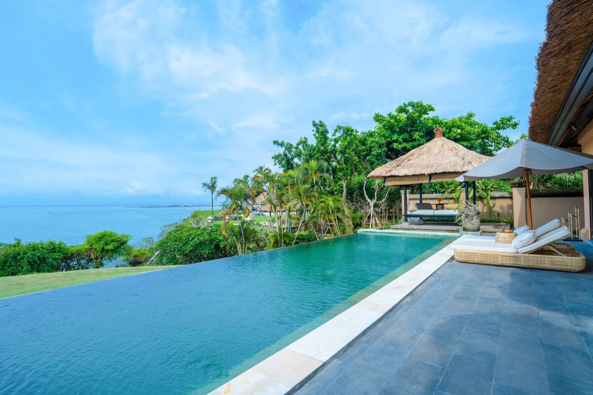 A pool with a view of the ocean, surrounded by lush greenery and a thatched-roof structure.