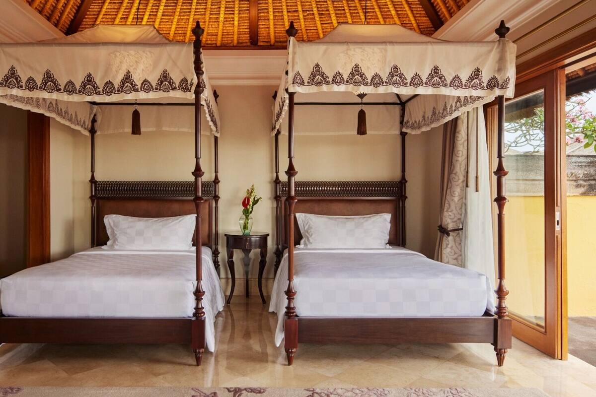 A room with two four-poster beds, a small table with a vase, and a view of greenery outside.