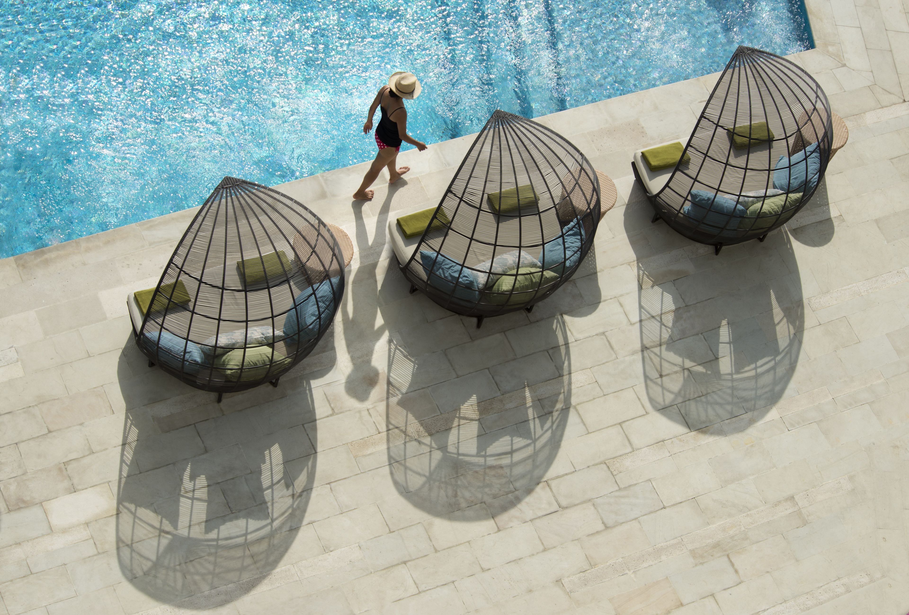 Uma área na beira da piscina com três espreguiçadeiras de metal, cada uma com almofadas, e uma pessoa passando.