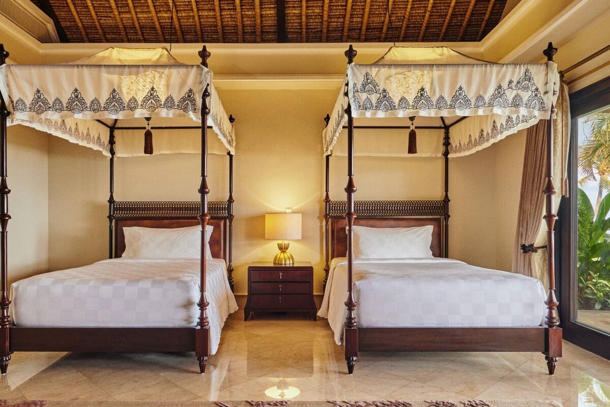 A four-poster bed with a canopy, a nightstand with a lamp, and a view of palm trees through a window.