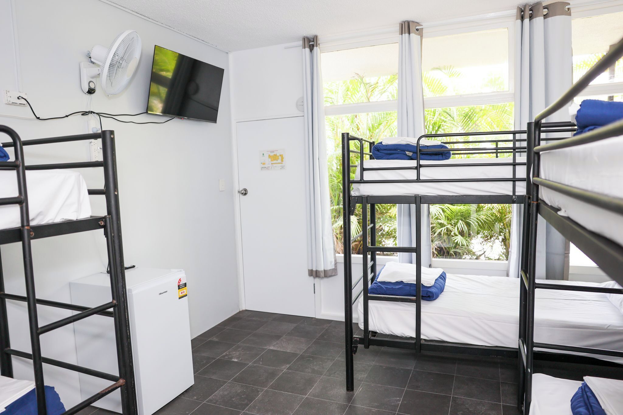 A dormitory room with bunk beds, a television, a fan, and a window with curtains.