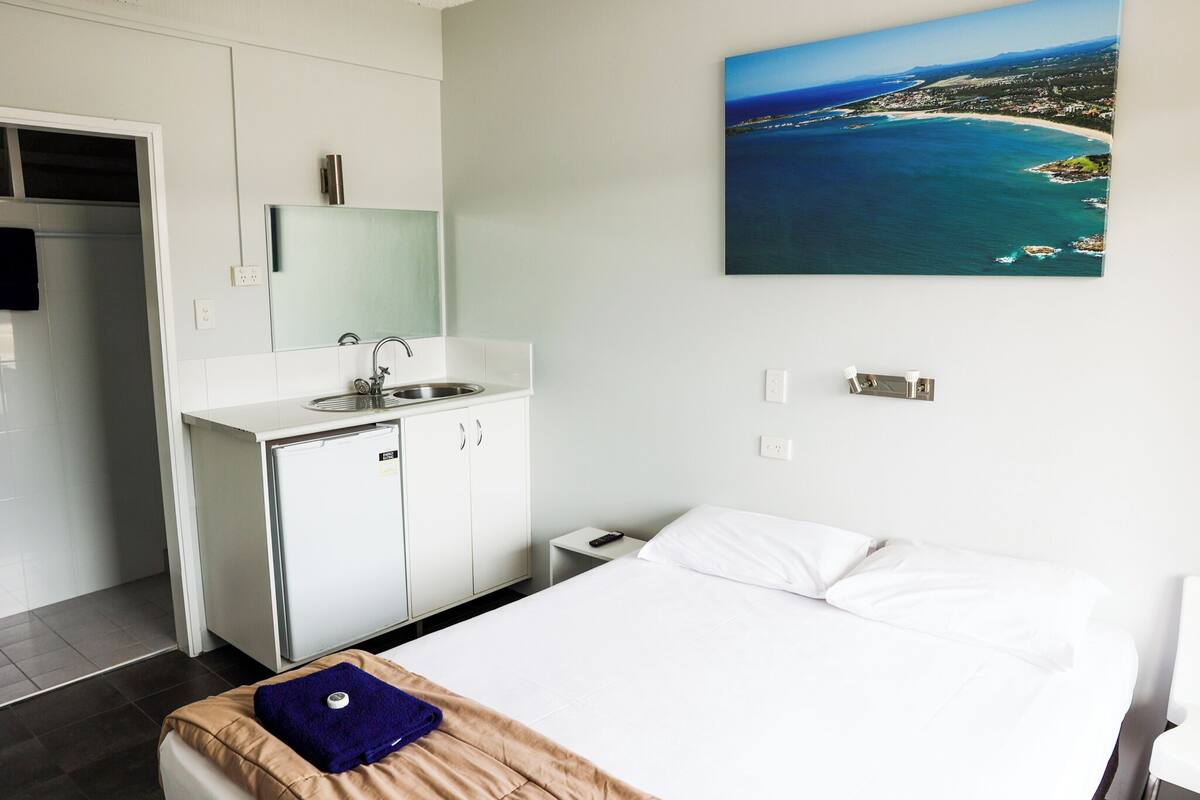 Habitación de hotel con cama, una kitchenette pequeña, baño y un cuadro de un paisaje costero.