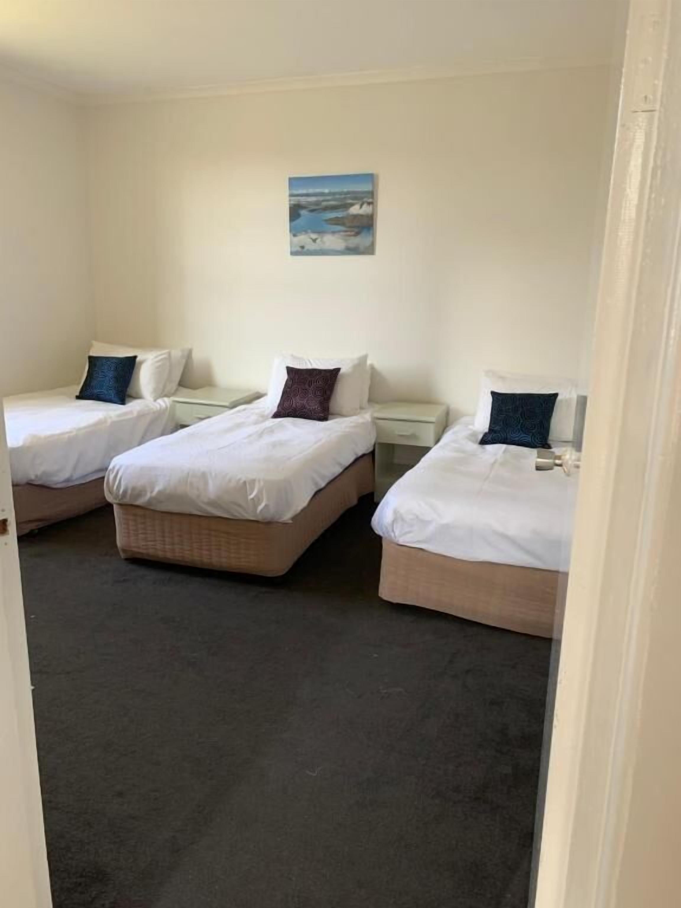 A room with three single beds, each with a white mattress and a pillow. There is a small table between the beds, and a painting on the wall above the middle bed.