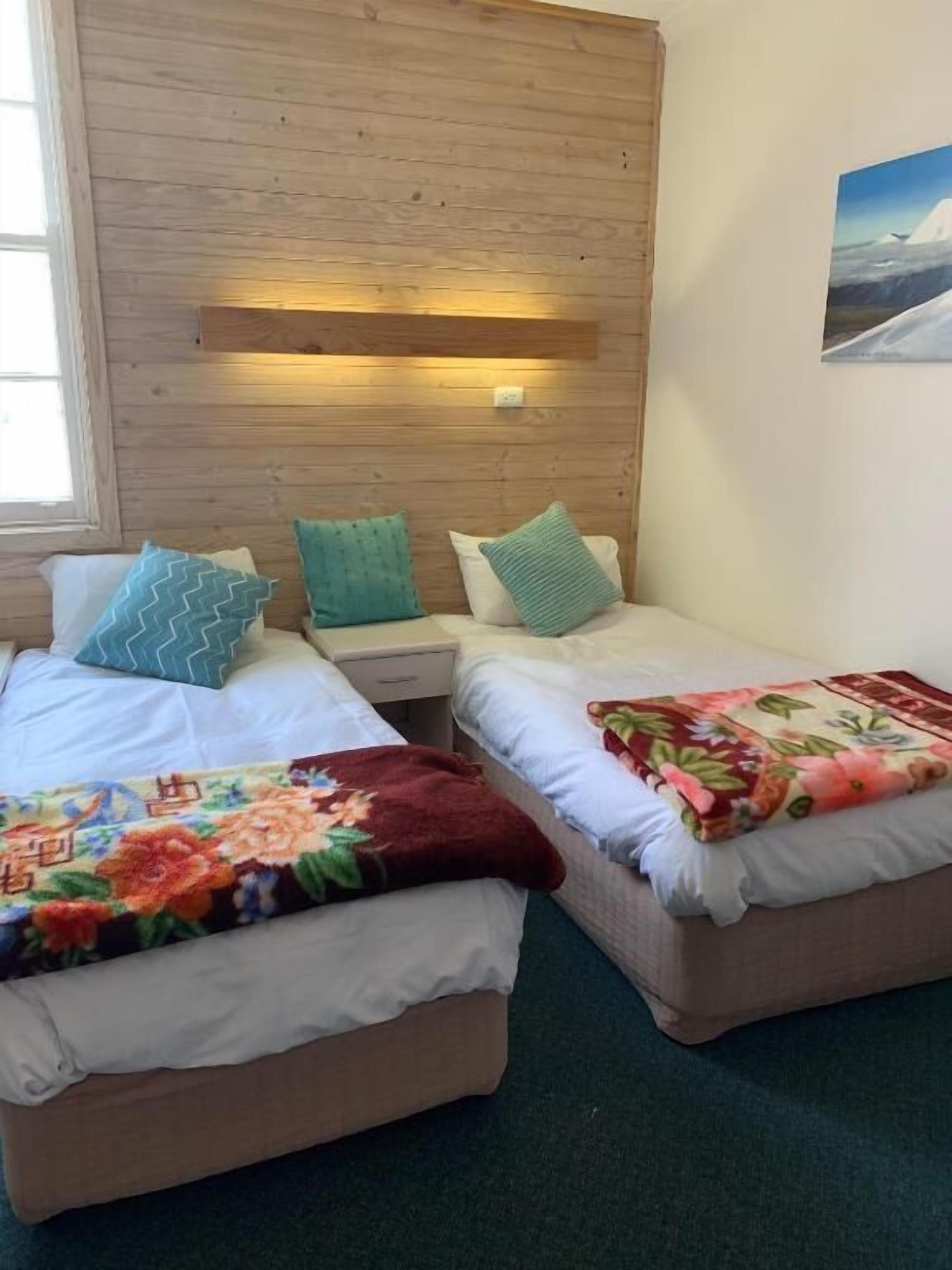 Una habitación con dos camas, cada una con una colcha estampada diferente. Hay un cabecero de madera con una lámpara sobre las camas y un cuadro en la pared a la derecha.