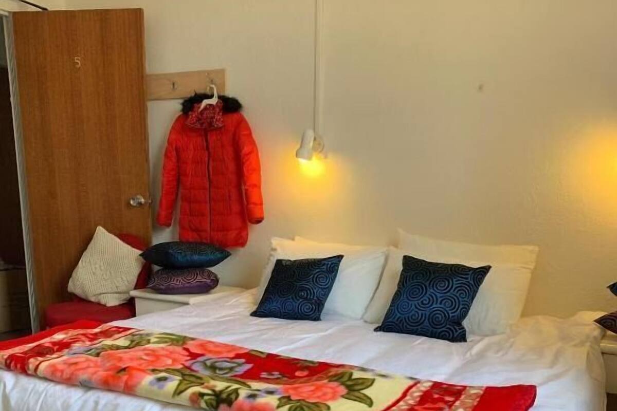 Una cama bien hecha con cubrecama floral, varios almohadones y un perchero con un abrigo rojo.