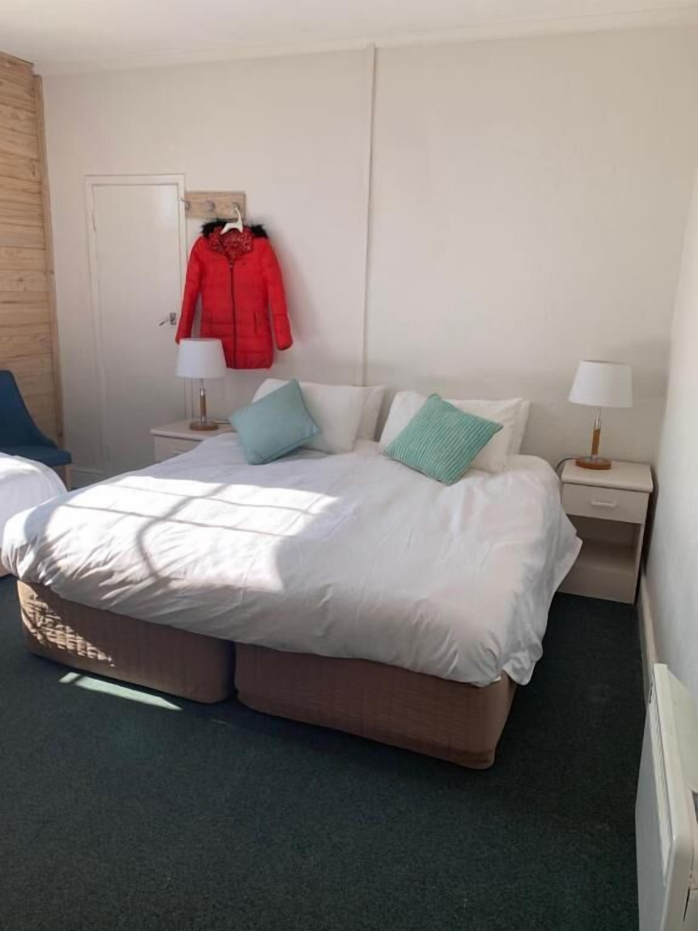Quarto de hotel com uma cama grande, duas mesas de cabeceira, uma cadeira e um cabideiro com um casaco vermelho.