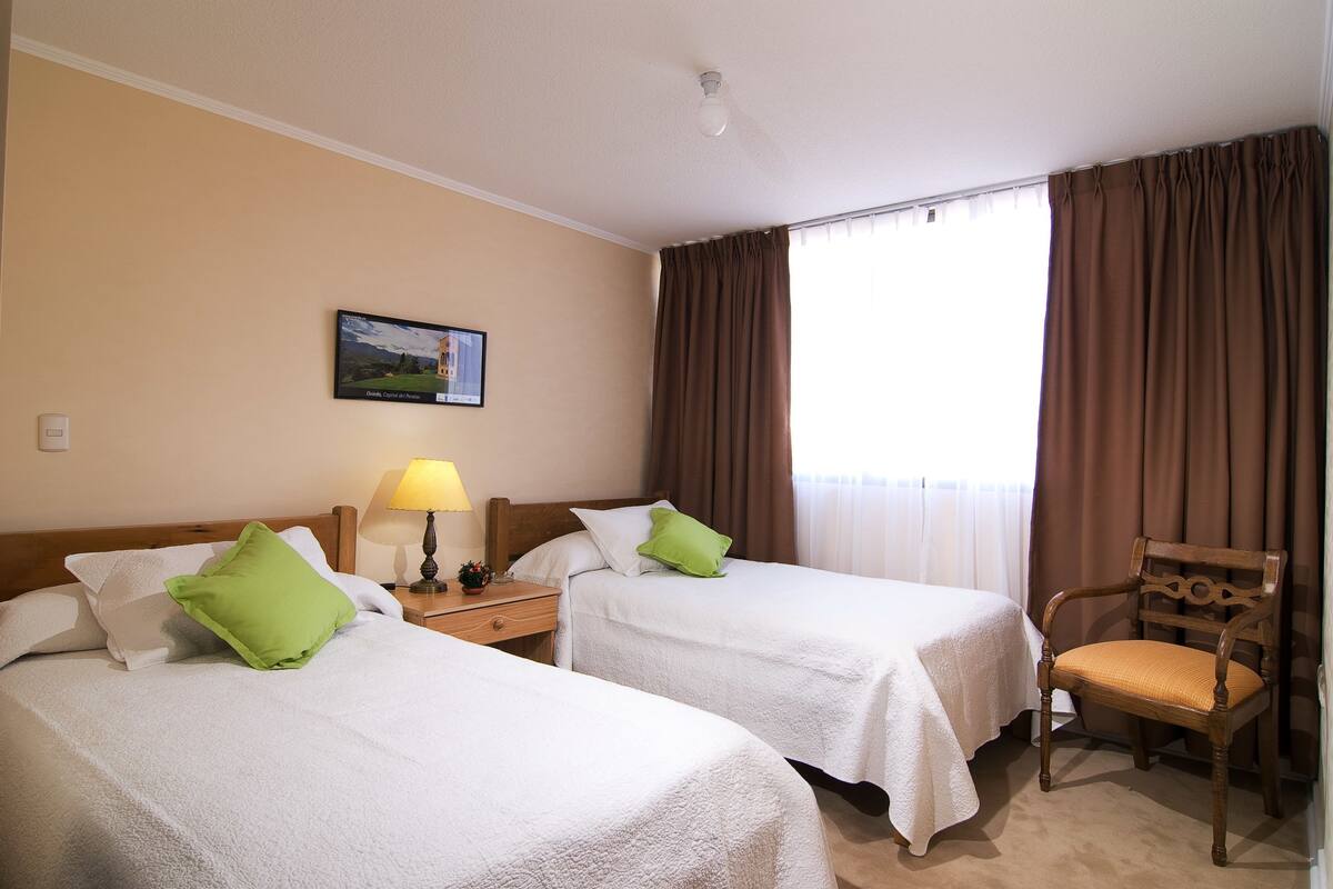 A hotel room with two beds, a chair, a nightstand with a lamp, and a painting on the wall.