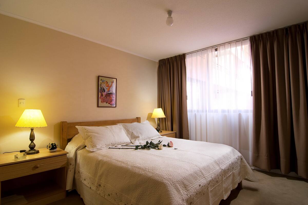 A neatly made bed with white linens, two bedside lamps, a small vase with flowers, and a painting on the wall.