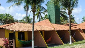 A row of triangular tents with a tiled roof, surrounded by palm trees and a building with a yellow facade. - Pousada Praia de Imbassaí (Mata de São João)