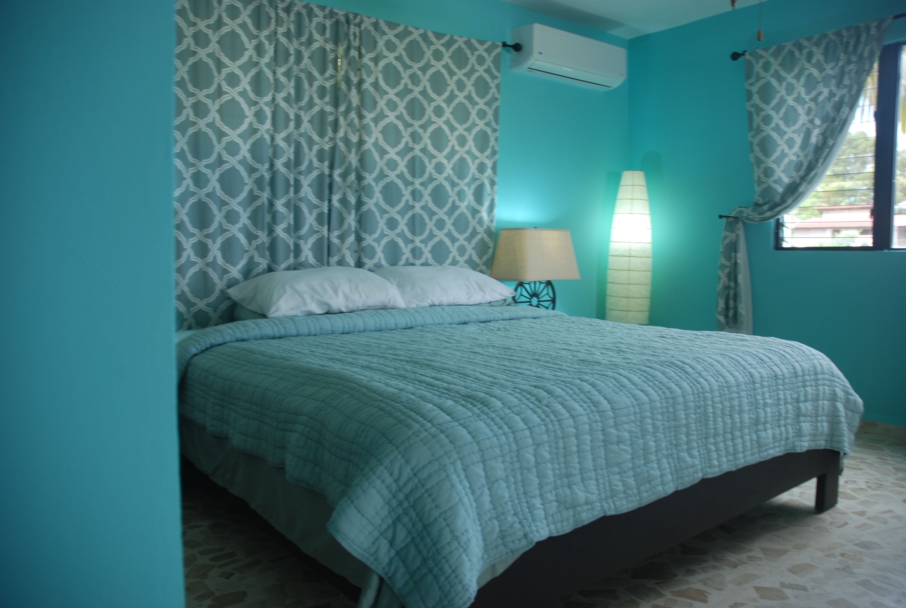 A bedroom with a bed, patterned curtains, a bedside lamp, and an air conditioning unit.