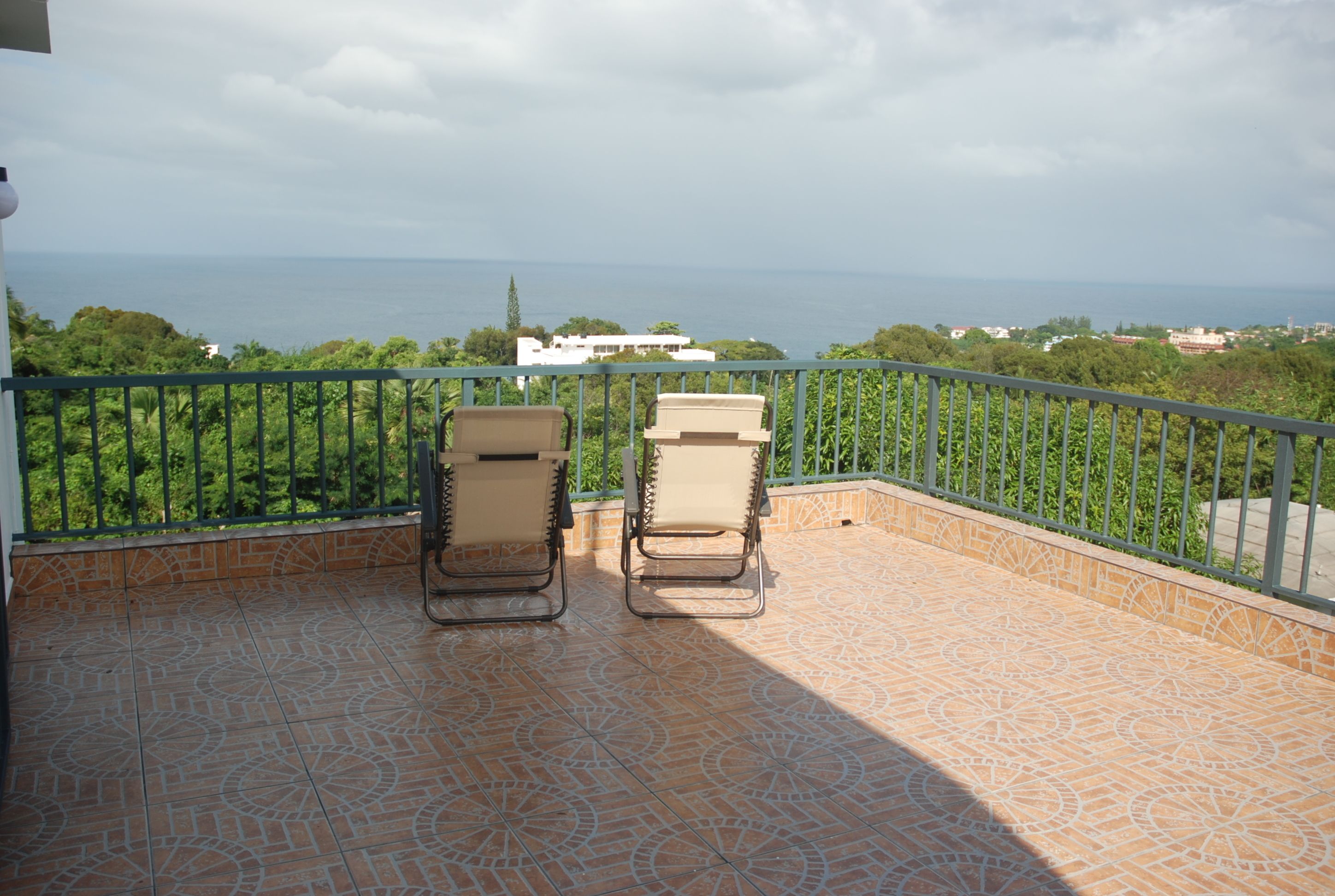 A balcony with two beige lounge chairs, a patterned tiled floor, and a view of the sea and greenery.