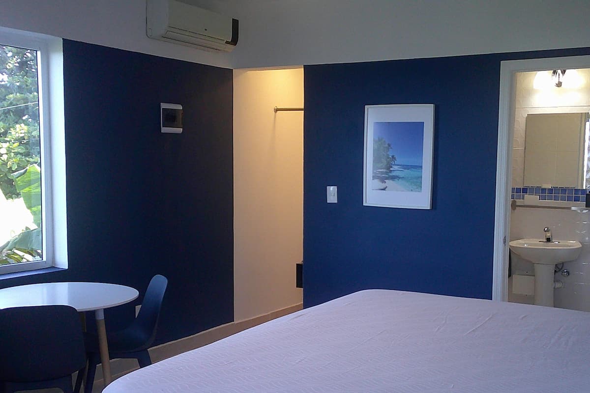 Een slaapkamer met een bed, een klein tafeltje, een stoel, een raam, een deur naar een badkamer en een foto in een lijst aan de muur.