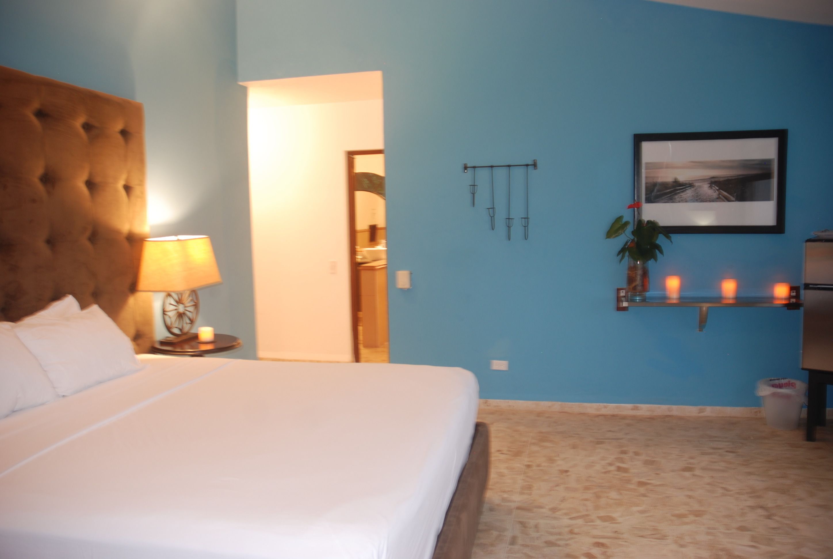Quarto com cabeceira estofada, luminária de cabeceira, quadro emoldurado e prateleira com velas.