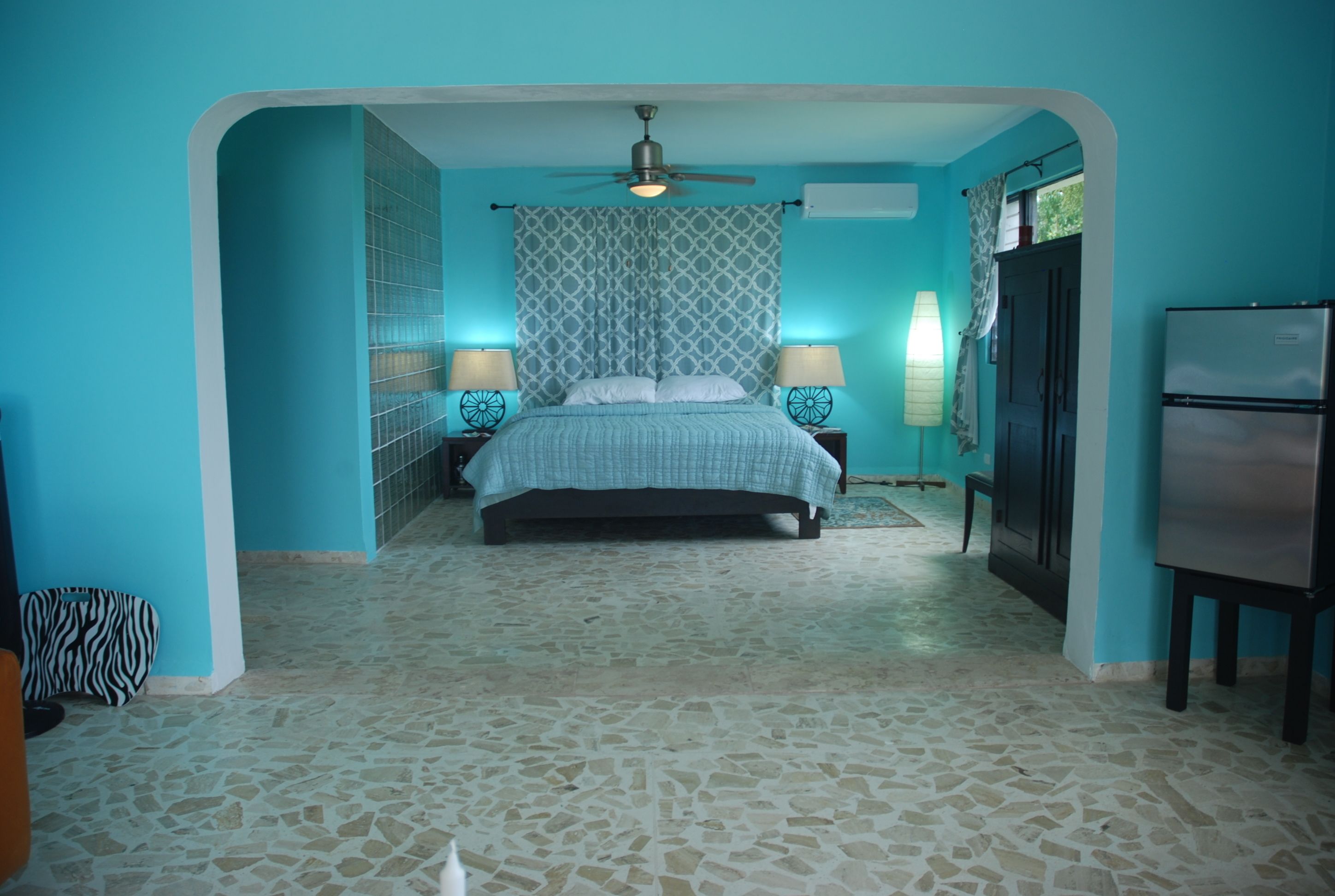 A bedroom with a bed, two bedside tables, a ceiling fan, a refrigerator, and a patterned wall.