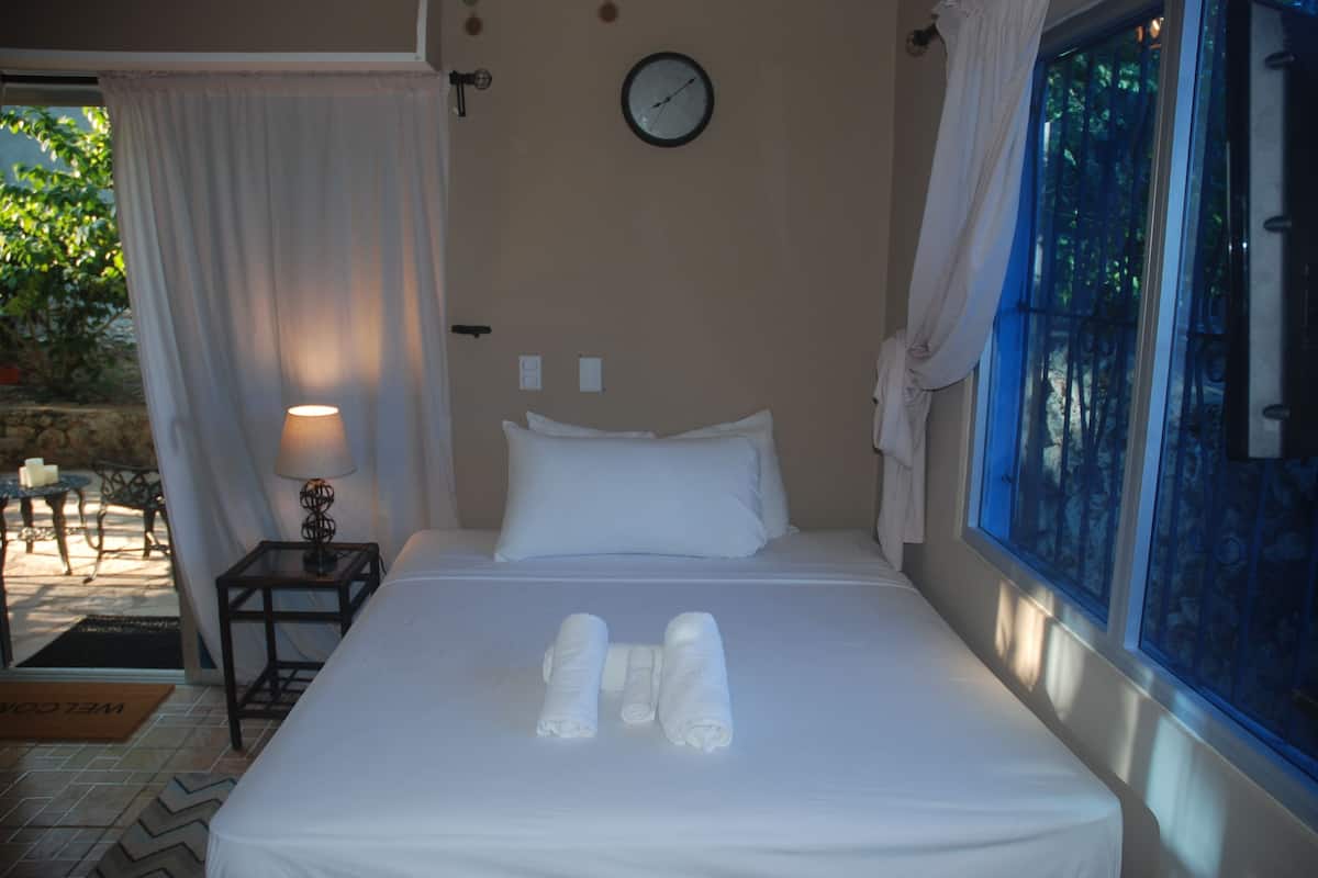 Een keurig opgemaakt bed met witte lakens en handdoeken, een nachtkastje met een lamp, een klok aan de muur en een raam met gordijnen.
