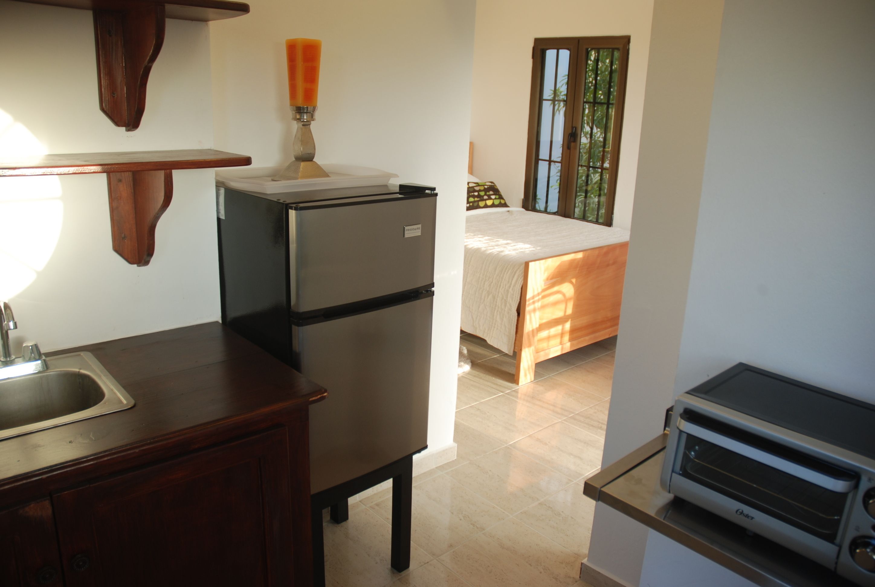 A compact kitchen with a refrigerator, a sink, and a toaster. A bed with a window is visible in the background.