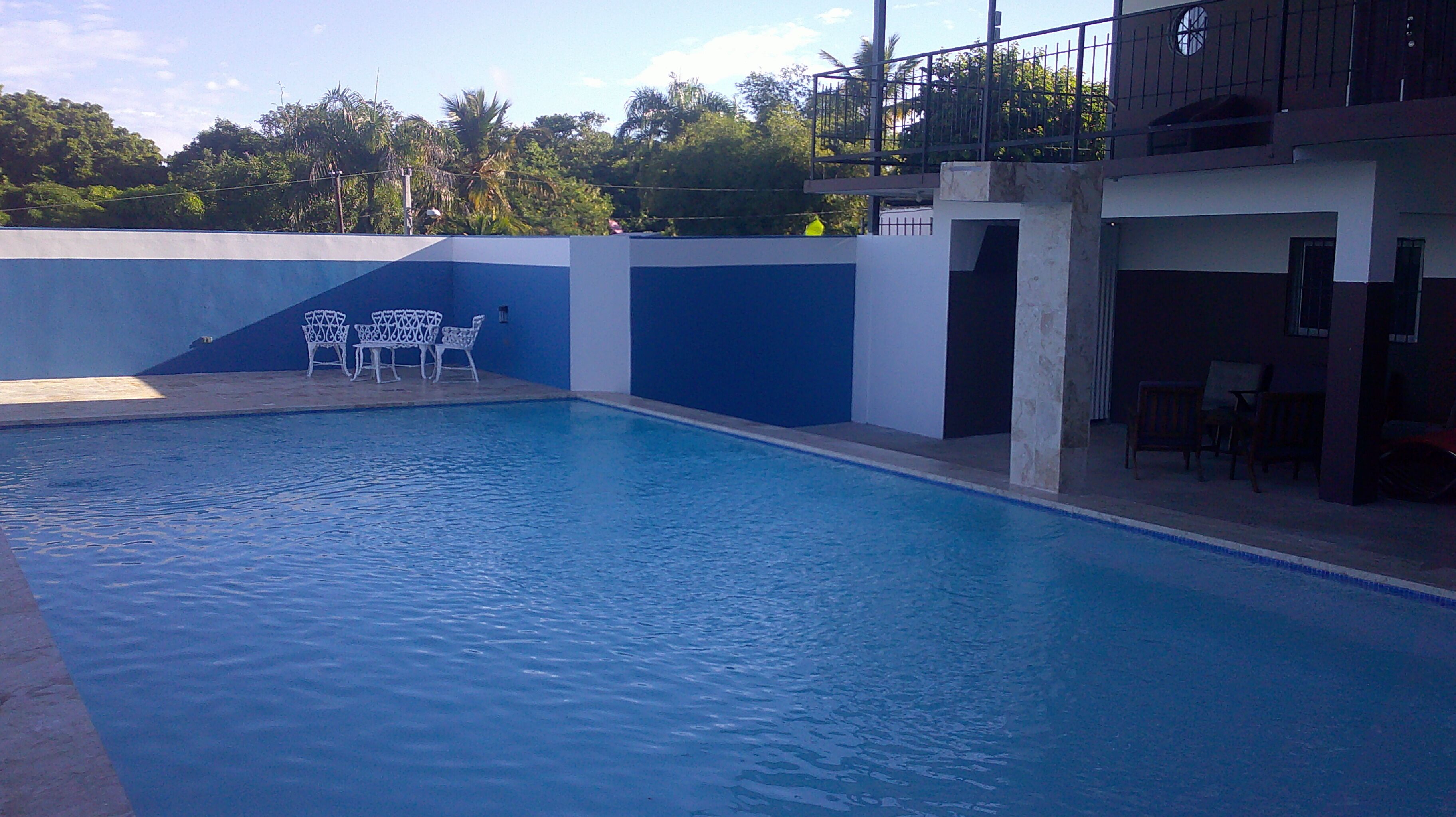 An empty swimming pool with a blue and white wall, a small seating area with white chairs, and a building with a balcony in the background.
