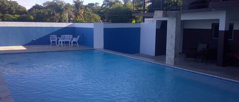 An empty swimming pool with a blue and white wall, a small seating area with white chairs, and a building with a balcony in the background.