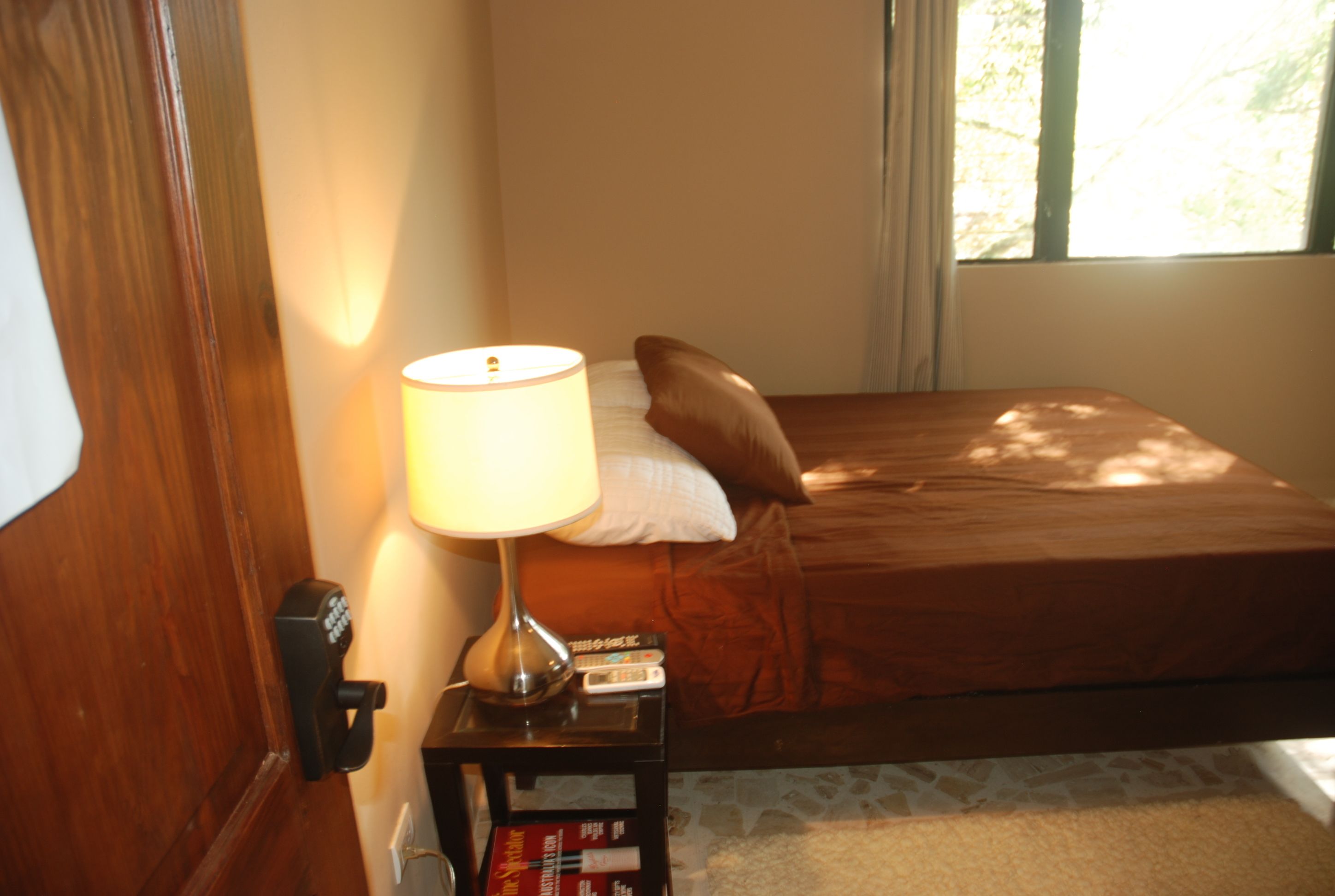 A bedroom with a bed, a bedside lamp, a nightstand with books, and a window with curtains.