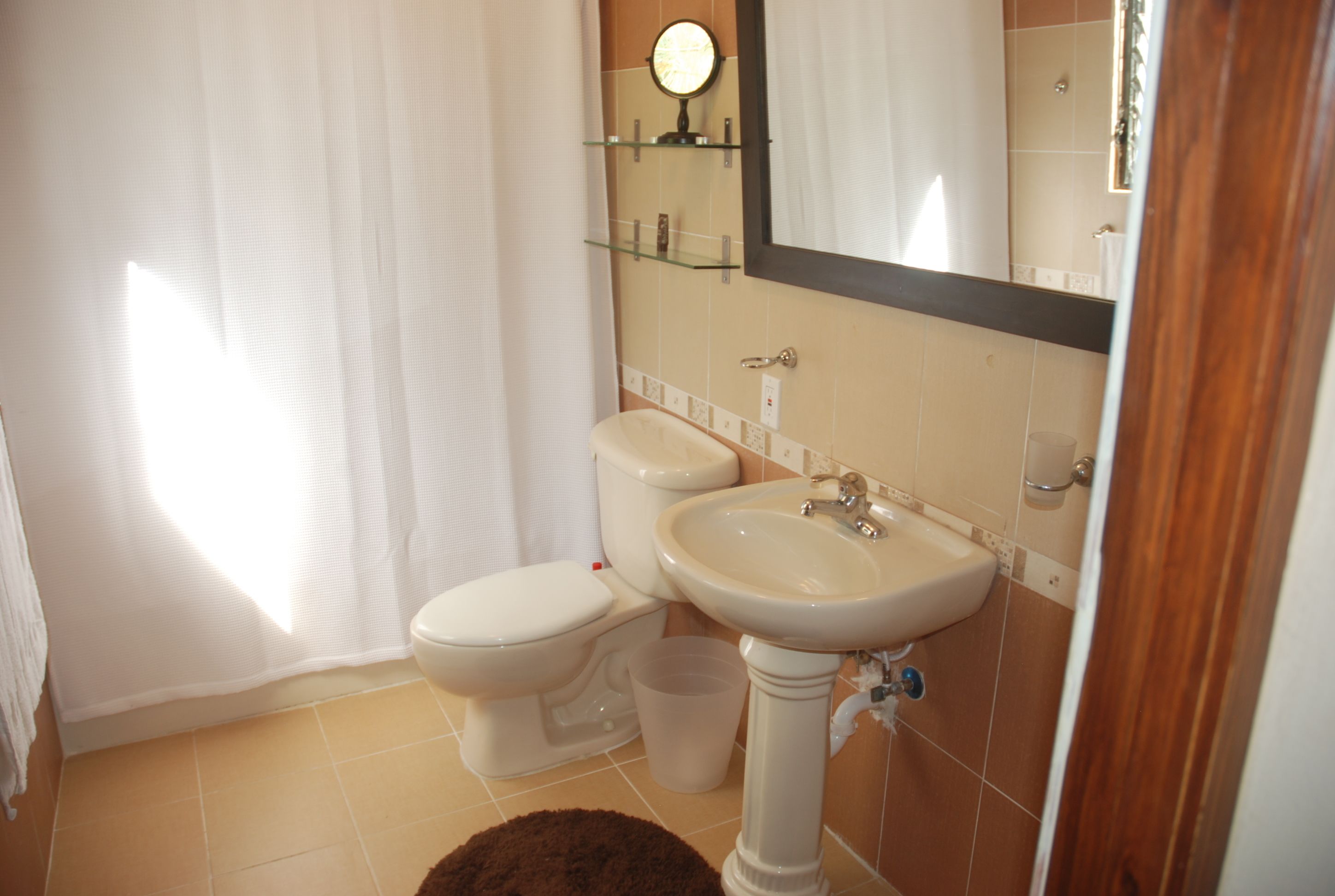 Un bagno con il water, un lavandino e uno specchio. Ci sono scaffali sopra lo specchio e una tenda per la doccia.