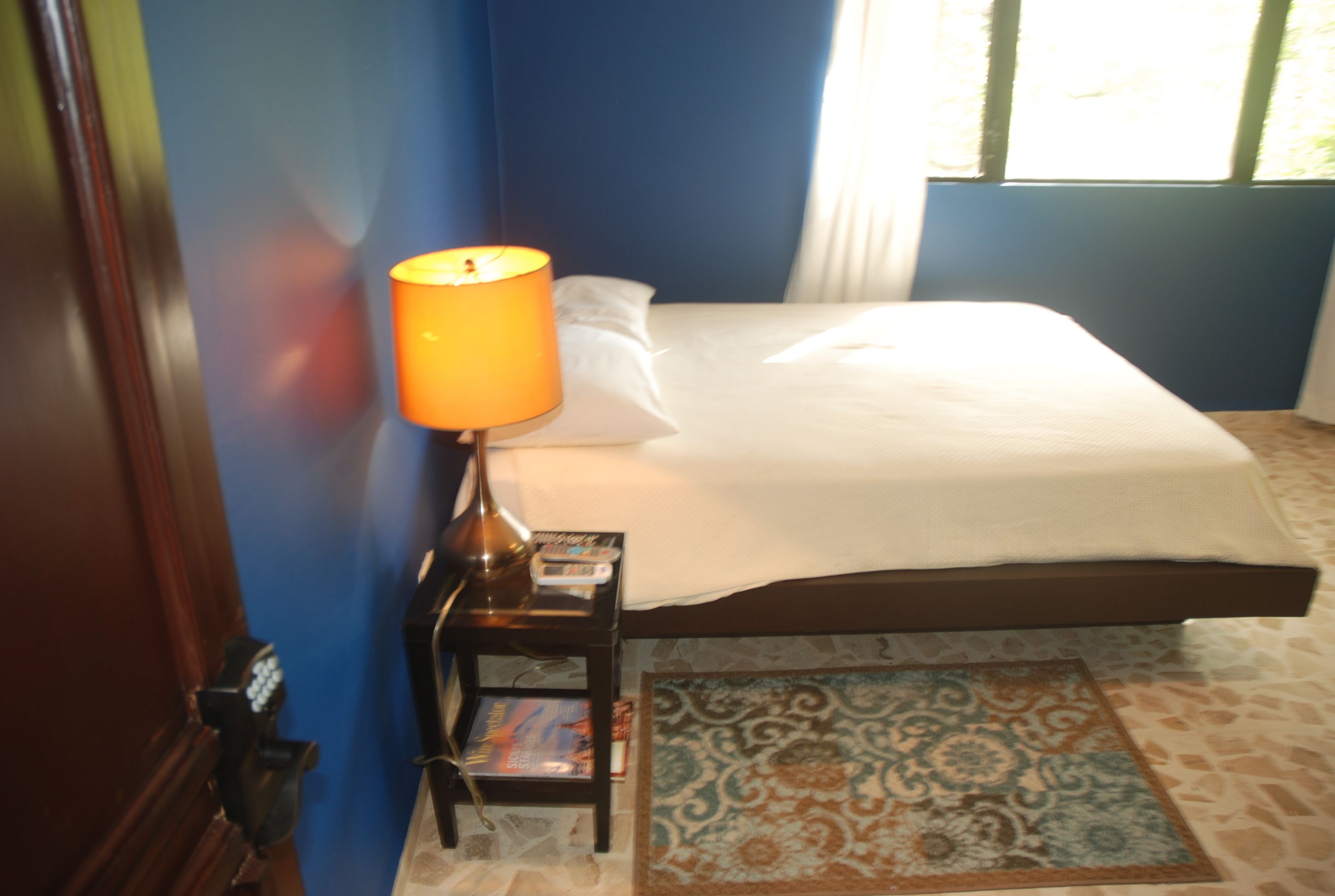 A neatly made bed with a white comforter and pillows, a small bedside table with a lamp and a magazine, and a patterned rug on the floor.