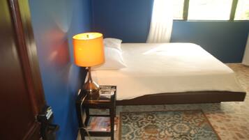 A neatly made bed with a white comforter and pillows, a small bedside table with a lamp and a magazine, and a patterned rug on the floor.