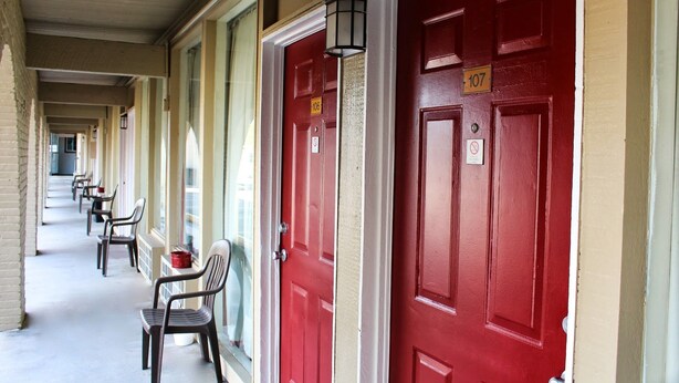 A row of red doors with numbered signs, a covered walkway, and outdoor chairs. - Executive Inn and Suites (Goliad)