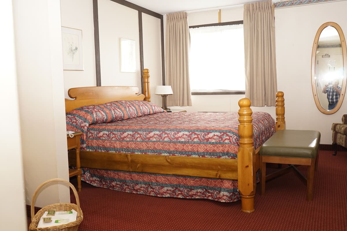Een hotelkamer met een houten bed, een stoel, een klein tafeltje, een spiegel en een raam met gordijnen.