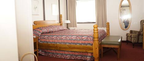A hotel room with a wooden bed, a green chair, a small table, a wicker basket, and a mirror on the wall.