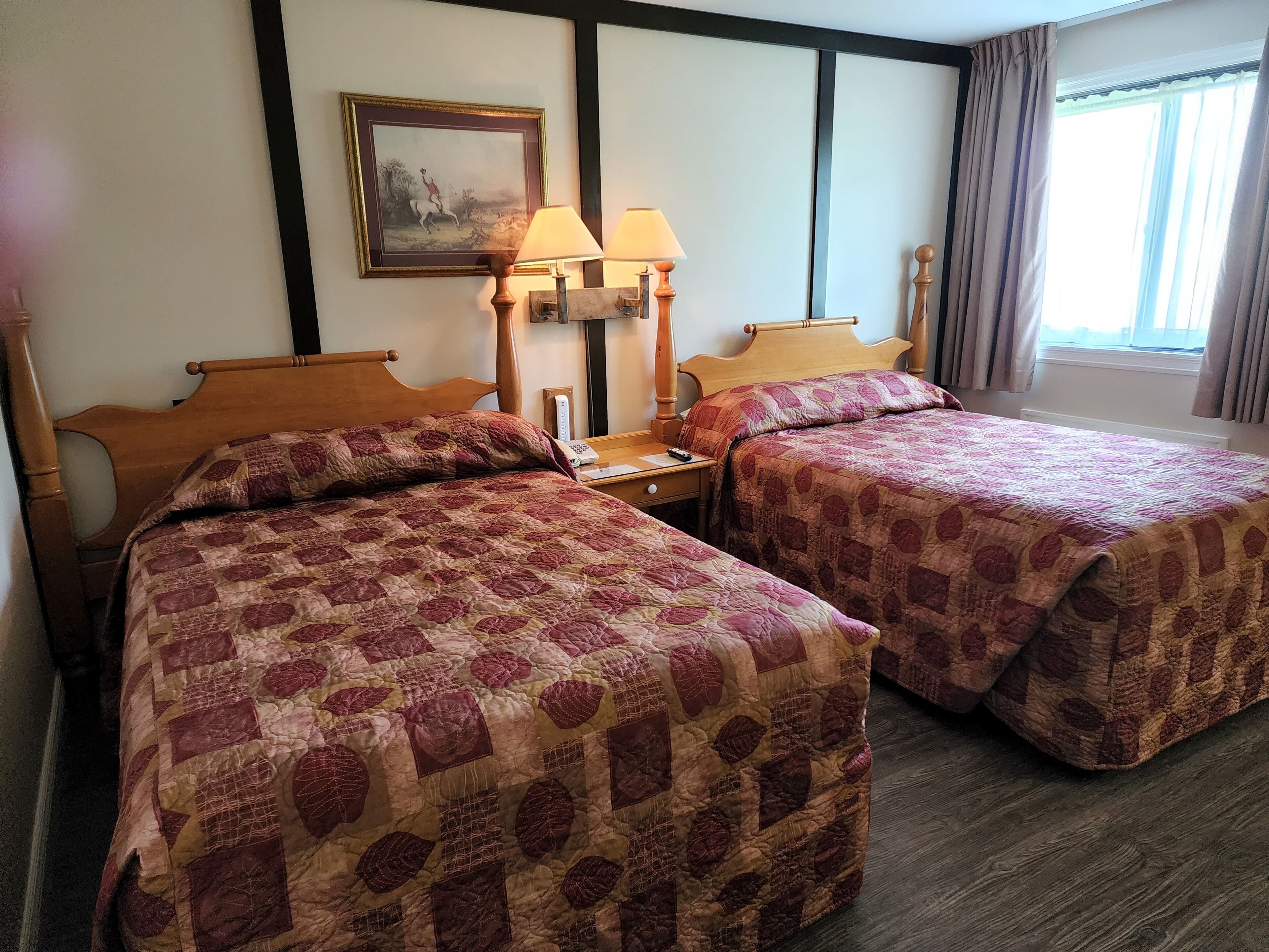 Une chambre d’hôtel avec deux lits, une table de chevet, une lampe, un tableau au mur et une fenêtre avec des rideaux.