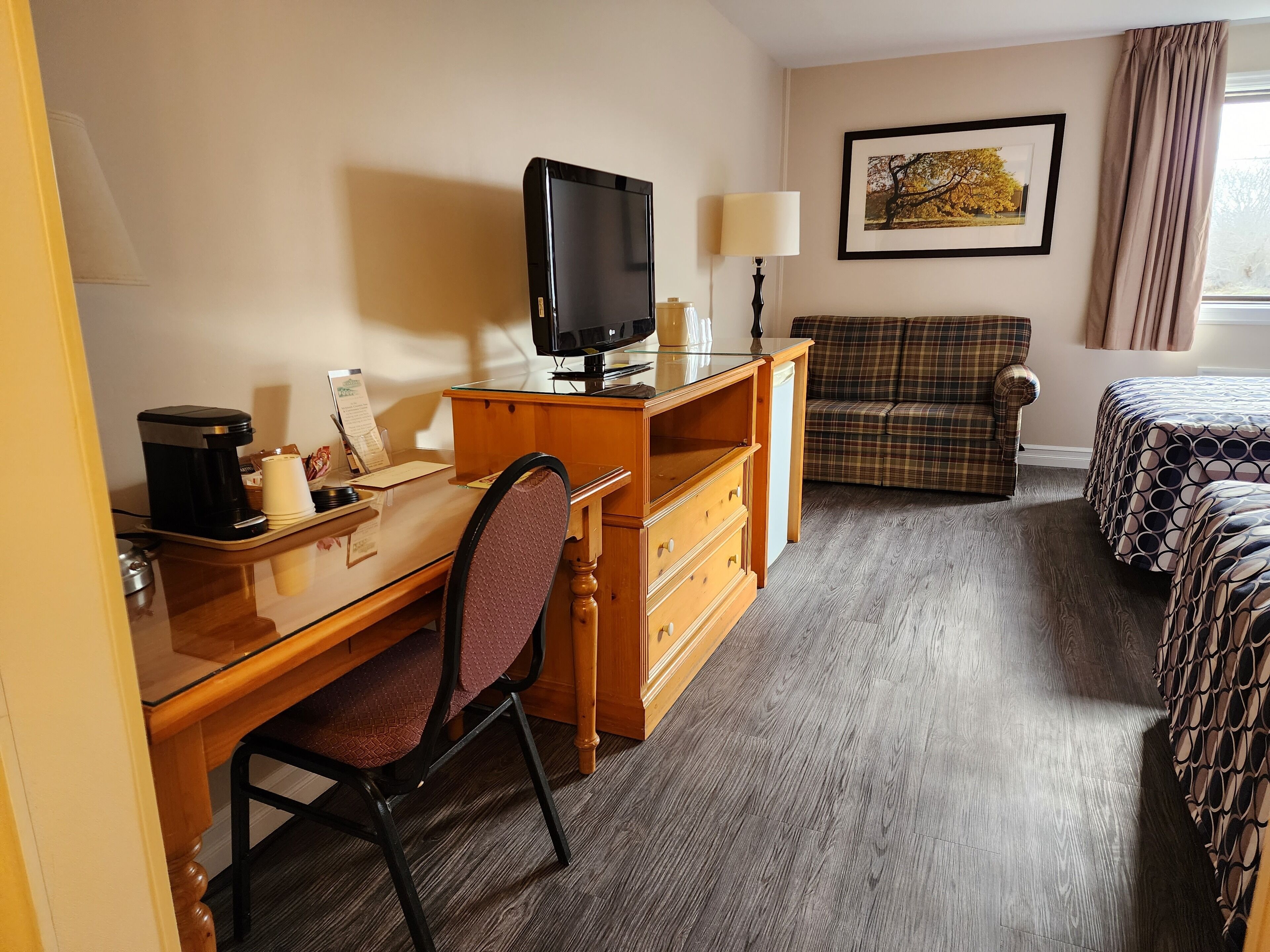 A hotel room with a wooden desk, a chair, a bed with patterned bedding, a plaid sofa, a television, and a framed picture on the wall.