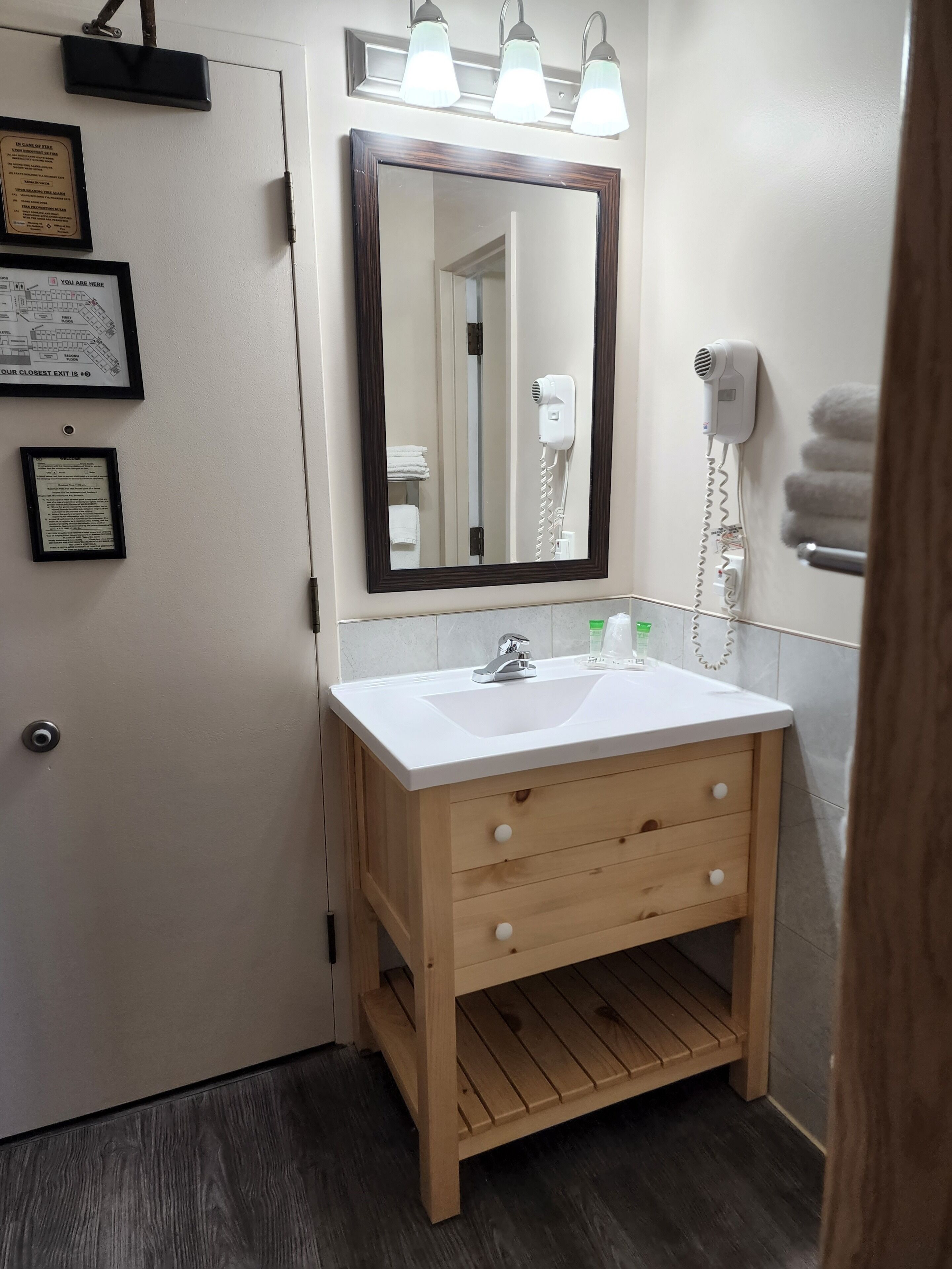 Une salle de bain avec un meuble-lavabo en bois, un grand miroir, un lavabo blanc et un sèche-cheveux fixé au mur.