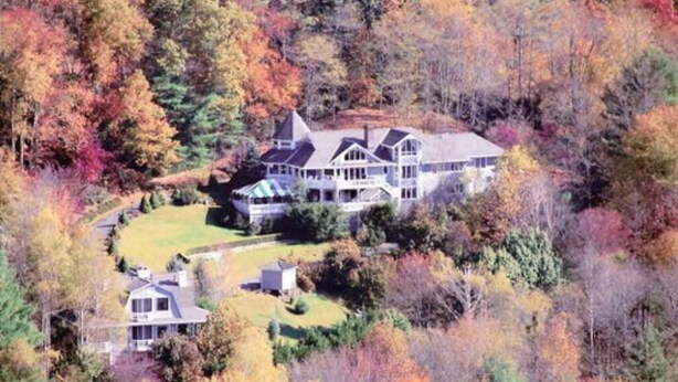 Een groot, twee verdiepingen hoog huis met een opvallende toren, omringd door een dicht bos met herfstkleuren.