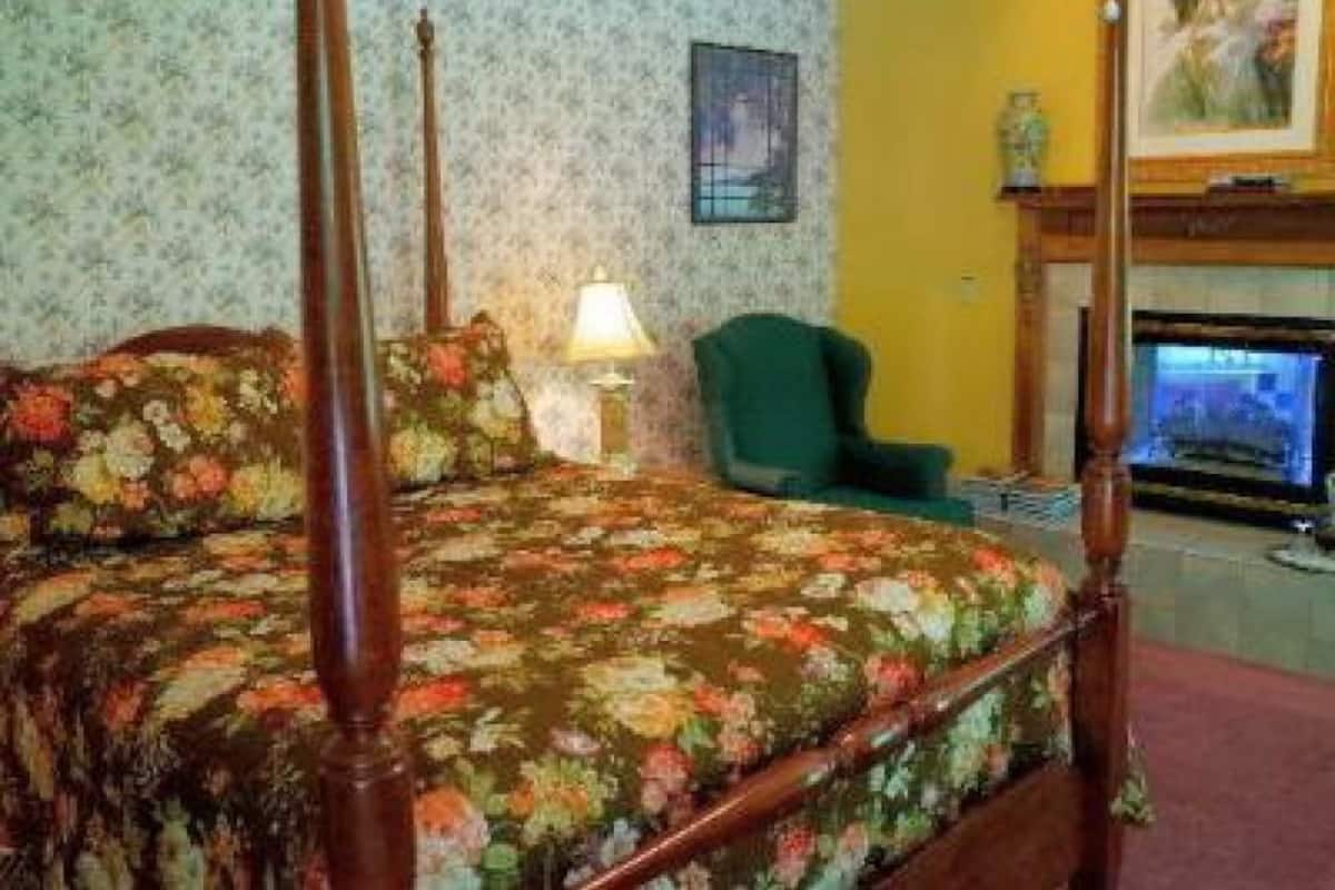 A four-poster bed with floral bedding, a green armchair, a lamp, and a fireplace with a painting above it.