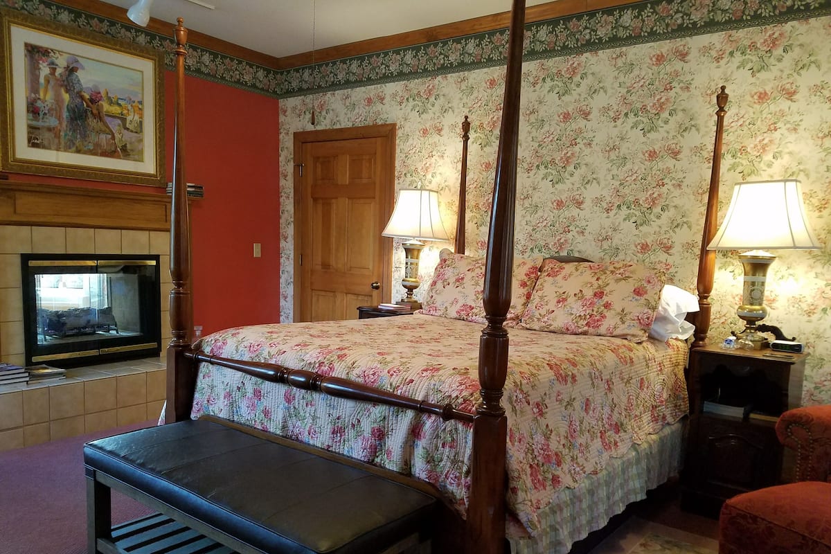 A bedroom with a four-poster bed, a bench, a fireplace, and floral wallpaper.