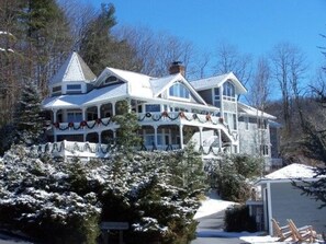 A large, multi-story house with a porch decorated with garlands, surrounded by snow-covered trees.