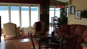A living room with a fireplace, a dining area, and large windows overlooking a body of water. - Pictured Rocks Bed And Breakfast (Munising)
